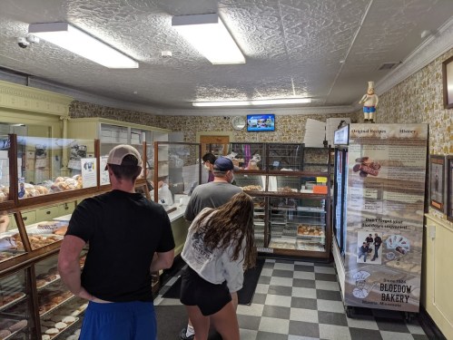 Customers line up in a bakery, looking at displays of baked goods, with a TV showing a program in the background.