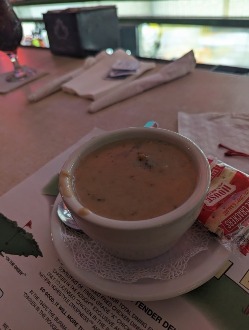 A small cup of creamy soup on a table, accompanied by a packet of crackers and a napkin.