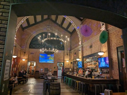 Interior of a lively restaurant with a bar, chandeliers, and colorful decor, featuring screens and patrons enjoying their meals.