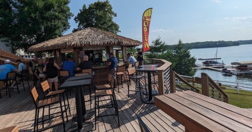 A lakeside bar with a thatched roof, outdoor seating, and patrons enjoying drinks on a sunny day.