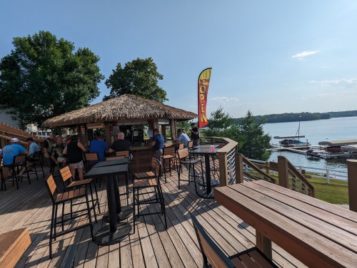A lakeside bar with a thatched roof, outdoor seating, and patrons enjoying the view on a sunny day.