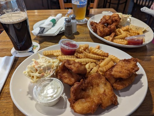 A plate of fried fish, crinkle-cut fries, coleslaw, and dipping sauces, with a glass of dark beer in the background.