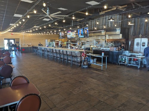 Interior of a restaurant featuring a long bar, wooden decor, and string lights, with tables and a kitchen area visible.