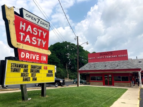 Sign for Hasty Tasty Pancake House with menu items listed, featuring a retro drive-in style building. Open 7 days.