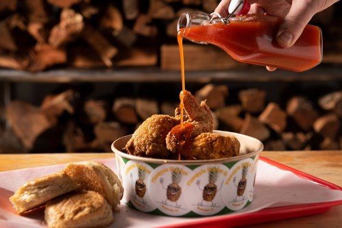 A hand pours hot sauce over crispy fried chicken in a bowl, with biscuits on the side and a rustic wooden backdrop.