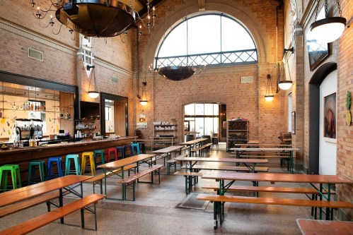 Interior of a spacious, modern hall with wooden benches, a bar, and large windows, featuring brick walls and industrial lighting.