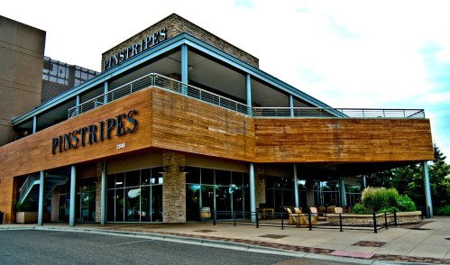 Exterior view of a modern building with wooden accents, featuring the sign "Pinstripes" and outdoor seating.