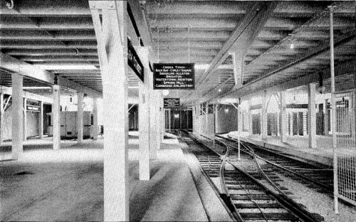 Black and white image of an empty train station platform with visible tracks and overhead signs.