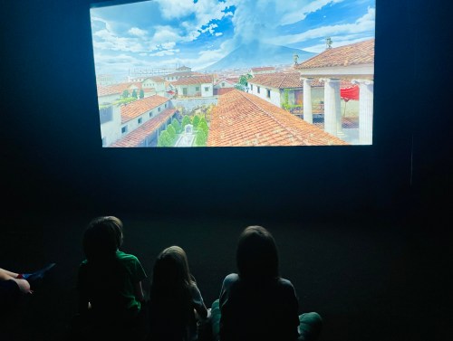 Three children sit in front of a large screen displaying a scenic view of rooftops and a blue sky.