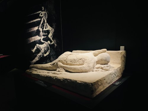 A sculpted figure lying on a stone slab, with a shadowy image of a person in the background.