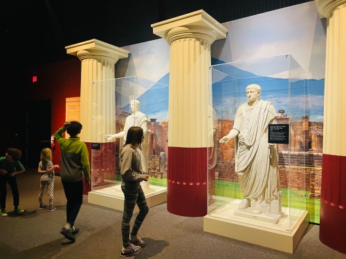 Children explore an exhibit featuring Roman statues in a museum, with columns and a scenic backdrop.