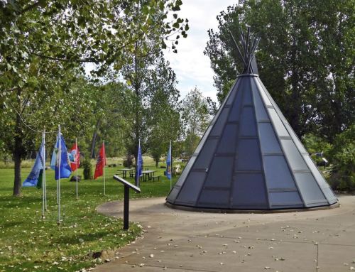 A modern teepee structure surrounded by trees and flags in a park setting.