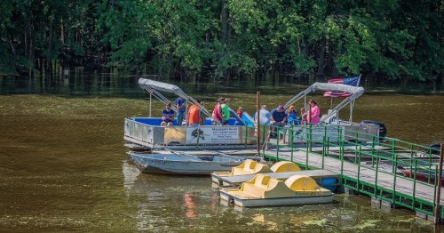 A group of people on a boat at a dock, surrounded by trees and calm water, with paddle boats nearby.