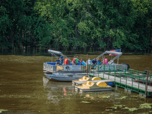 Two boats docked at a green pier on a calm lake, with people enjoying the sunny day surrounded by lush trees.