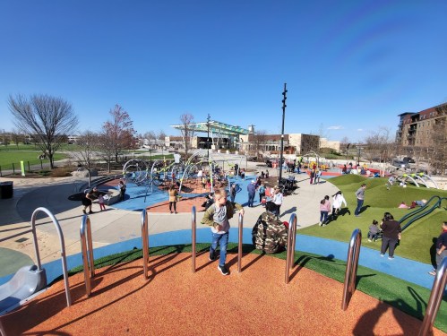 A vibrant playground filled with children playing, swings, and families enjoying a sunny day. Green grass and blue sky visible.
