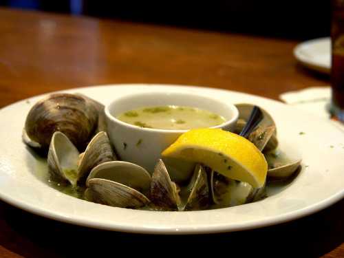 A bowl of clam chowder surrounded by fresh clams and a lemon wedge on a wooden table.