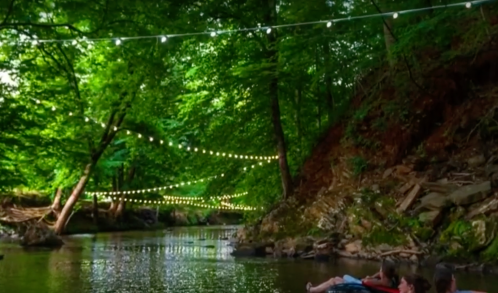 A serene river scene with string lights above, surrounded by lush greenery and people relaxing by the water.