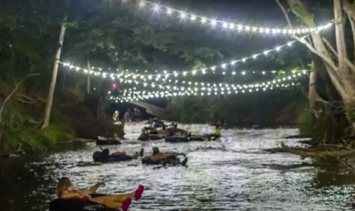 A serene night scene of people floating on inner tubes down a river, illuminated by string lights above.
