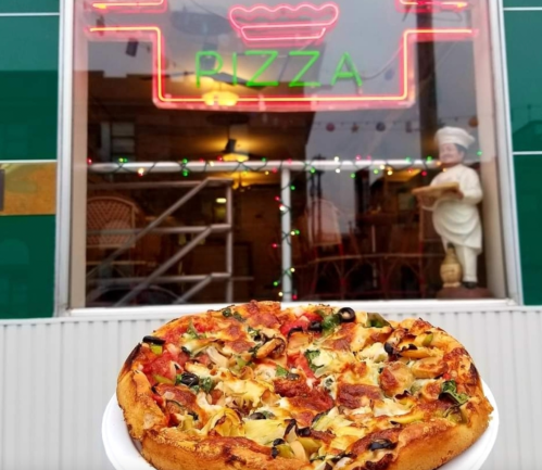 A freshly baked pizza with various toppings in front of a pizzeria with a neon "PIZZA" sign.