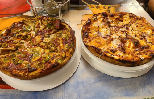 Two pizzas on white plates: one topped with green olives and vegetables, the other with cheese and meats.