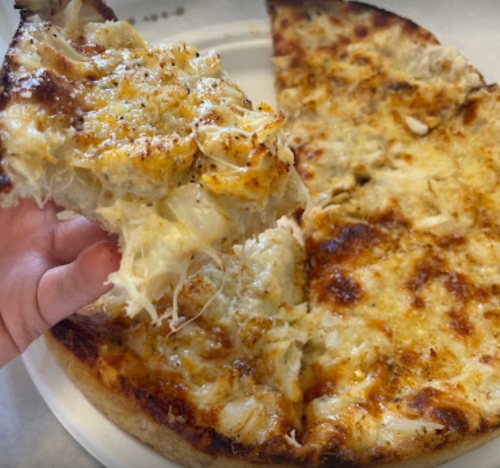 A hand holding a slice of cheesy pizza, with a golden crust, next to a partially eaten pizza on a plate.