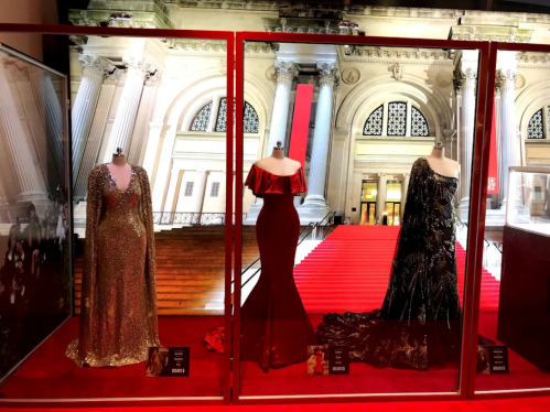Three elegant dresses displayed in glass cases, set against a backdrop of a grand staircase and red carpet.