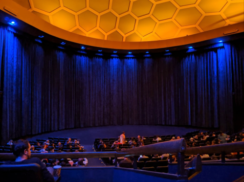 A dimly lit theater with blue curtains and a honeycomb ceiling, filled with seated audience members.