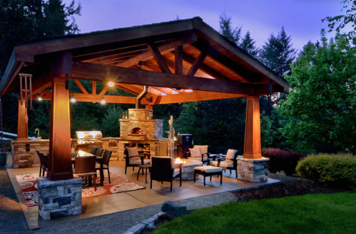 A cozy outdoor patio with a stone fireplace, seating area, and grill, surrounded by trees at dusk.