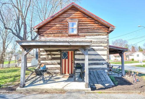 A rustic log cabin with a porch, featuring a door decorated with an American flag, surrounded by greenery.