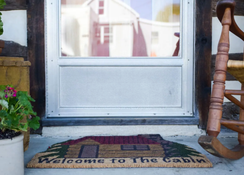 A welcome mat with a cabin design placed at the entrance of a home, next to a potted plant and a rocking chair.