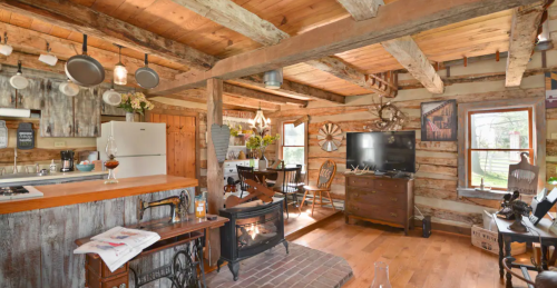 Cozy rustic cabin interior with wooden beams, a fireplace, kitchen area, and dining space. Warm and inviting atmosphere.