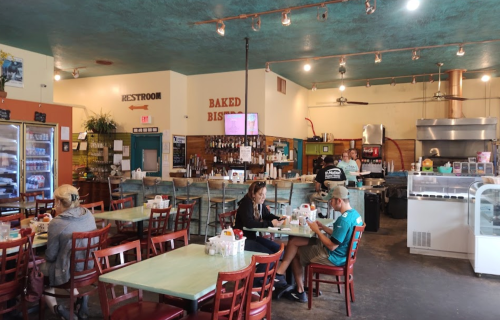 A cozy café interior with wooden tables, chairs, and a visible kitchen area. Customers enjoy their meals.