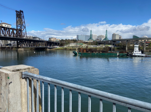 A view of a river with a barge, bridges, and city buildings under a blue sky with scattered clouds.