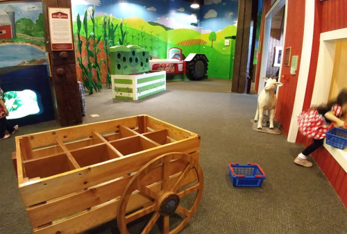 A colorful indoor play area featuring a wooden cart, tractor, cow statue, and vibrant farm-themed murals.