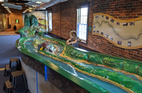 A child interacts with a colorful topographical model of a landscape in a brick-walled educational space.