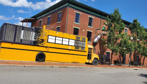 A bright yellow food truck parked beside a brick building with trees and blue sky in the background.