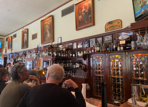 Two patrons sit at a bar with a wooden counter, surrounded by framed artwork and shelves of bottles.