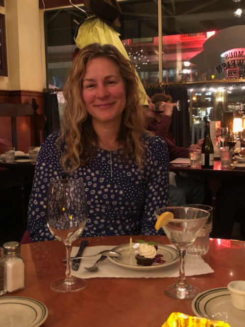 A woman with curly hair smiles at a table set with a dessert and drinks in a cozy restaurant setting.
