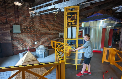 A child operates a yellow crane in a hands-on science exhibit with brick walls and various structures.