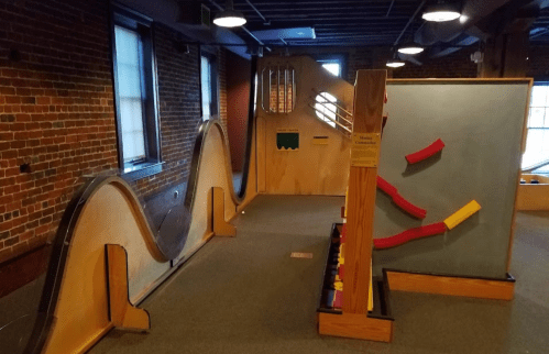 A playful indoor exhibit featuring curved ramps and colorful interactive panels in a brick-walled space.