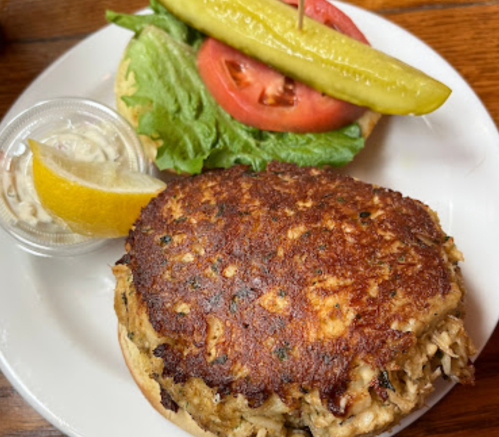 A large crab cake on a plate with lettuce, tomato, pickle, a lemon wedge, and a side of coleslaw.