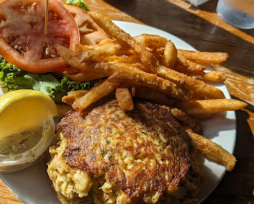 A plate featuring a crab cake, crispy fries, lettuce, tomato slices, and a lemon wedge.