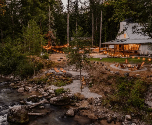 A serene riverside scene featuring a cozy cabin, outdoor seating, and string lights among lush trees at dusk.