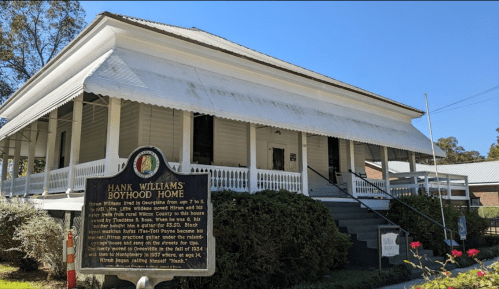 Historic Hank Williams Boyhood Home, a white wooden house with a porch, featuring a commemorative plaque in front.