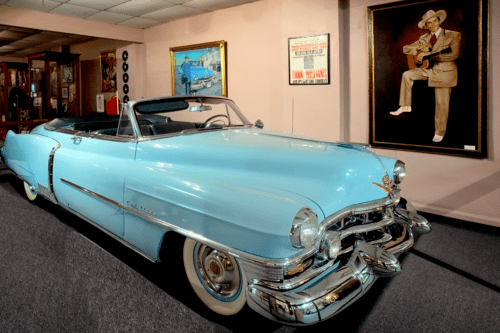 A vintage blue convertible car displayed in a museum, with framed portraits on the wall behind it.