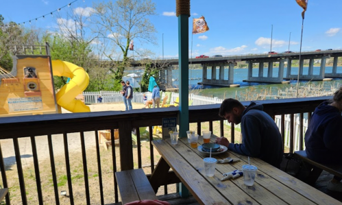 A person sits at a wooden table eating, with a playground and a bridge visible in the background by the water.