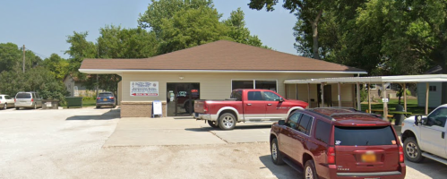 A small building with a brown roof, featuring a sign, surrounded by parked cars in a grassy area.