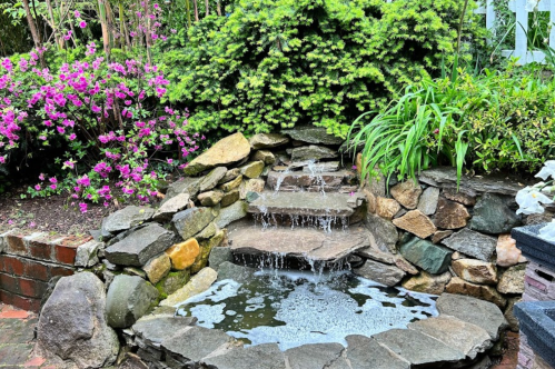 A serene garden scene featuring a small stone waterfall surrounded by lush greenery and vibrant pink flowers.