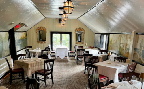 Cozy restaurant interior with elegant tables, wooden chairs, and large windows, featuring soft lighting and decorative mirrors.