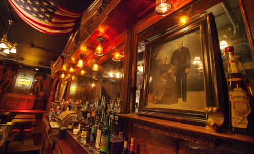 A vintage bar scene featuring a portrait, illuminated by warm lights and decorated with an American flag.